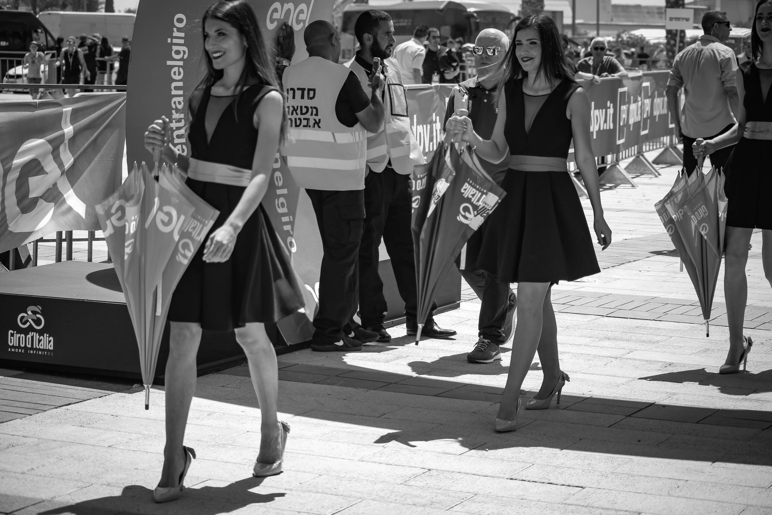 Podium hosts walking through hard midday sun at a Giro d’Italia stage start, black and white