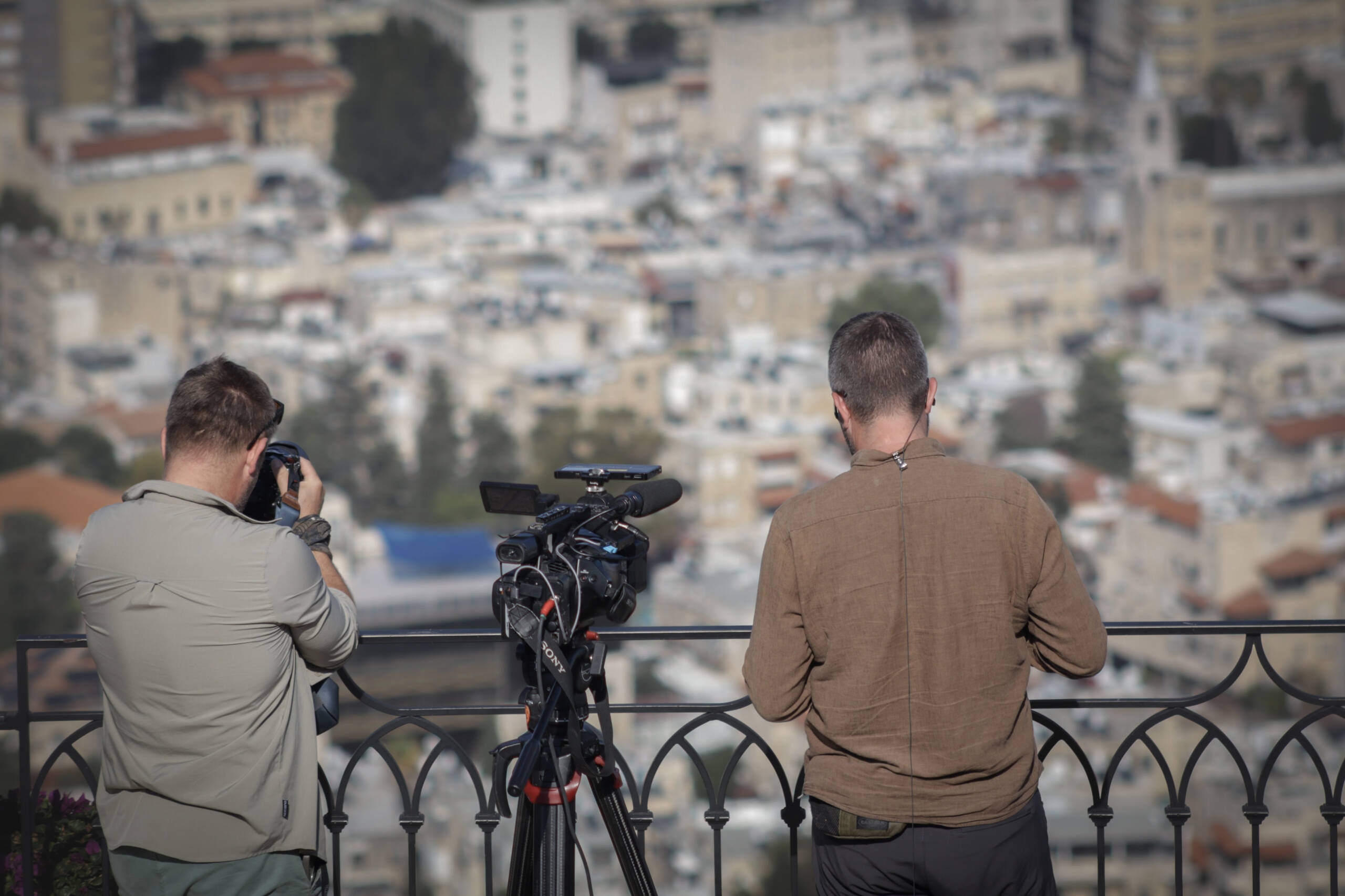 Two men filming a city from a balcony, one with a handheld camera and one beside a tripod-mounted Sony video rig