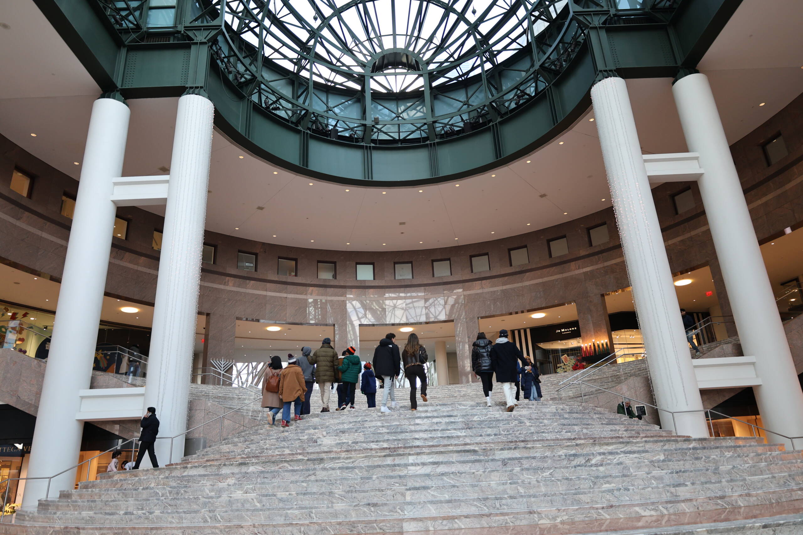 Shoppers climbing a wide marble staircase inside a luxury shopping centre atrium, beneath a circular glass and steel dome