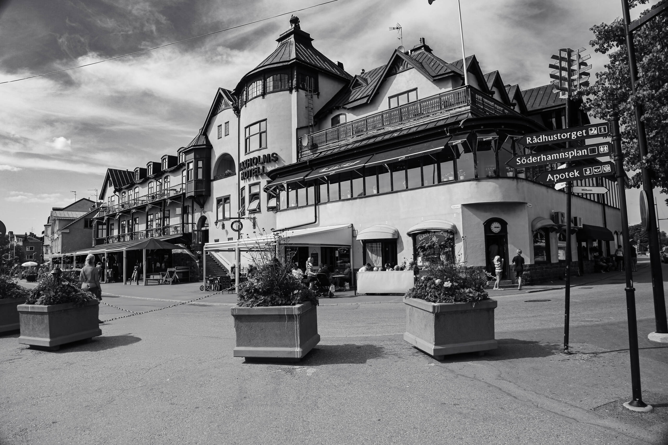Vaxholms Hotell photographed from the street corner in black and white, hard summer sunlight, concrete planters in the foreground, direction signs reading Rådhustorget and Söderhamnsplan