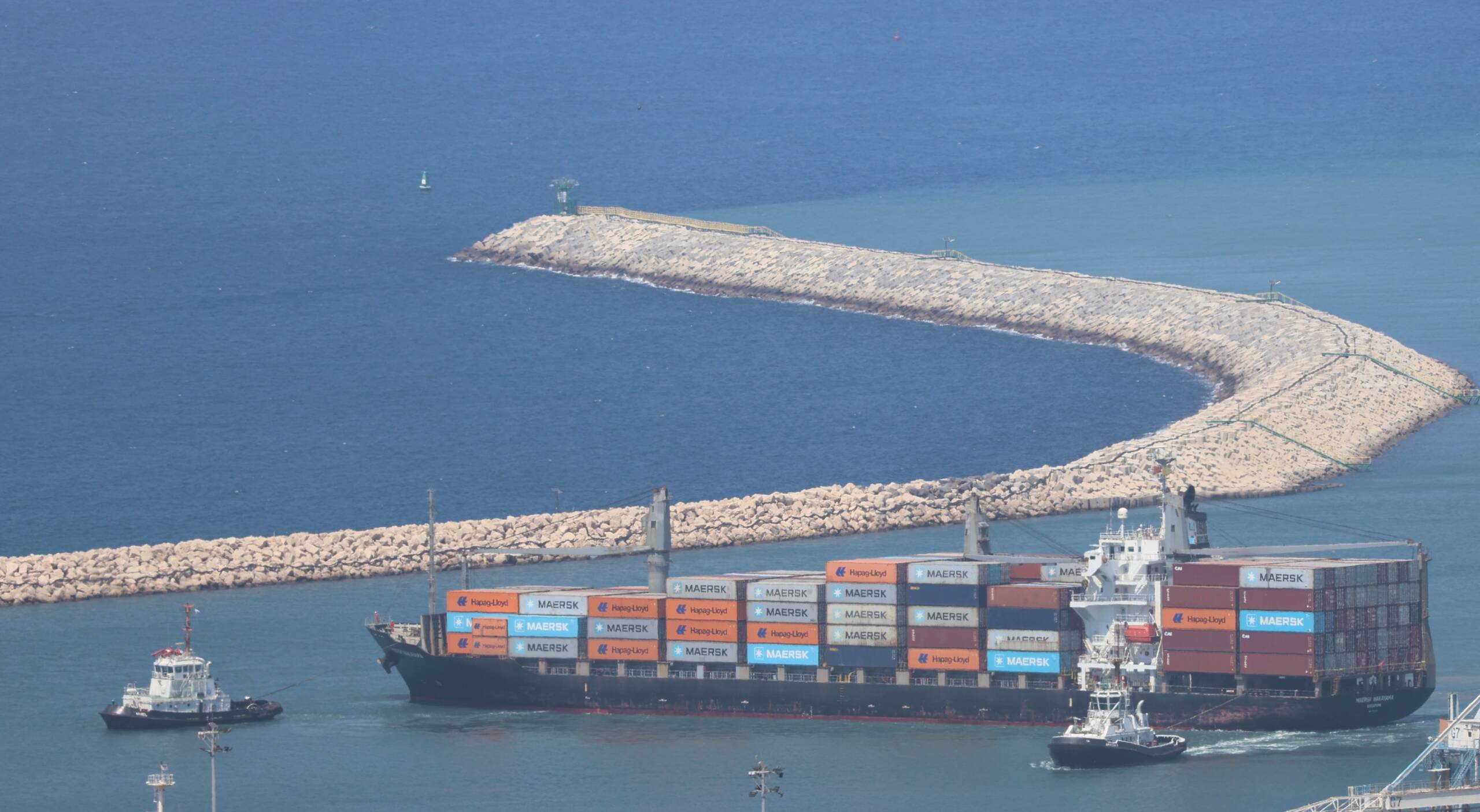 A geared container ship under tug escort passing the curved rubble-mound breakwater at a harbour entrance