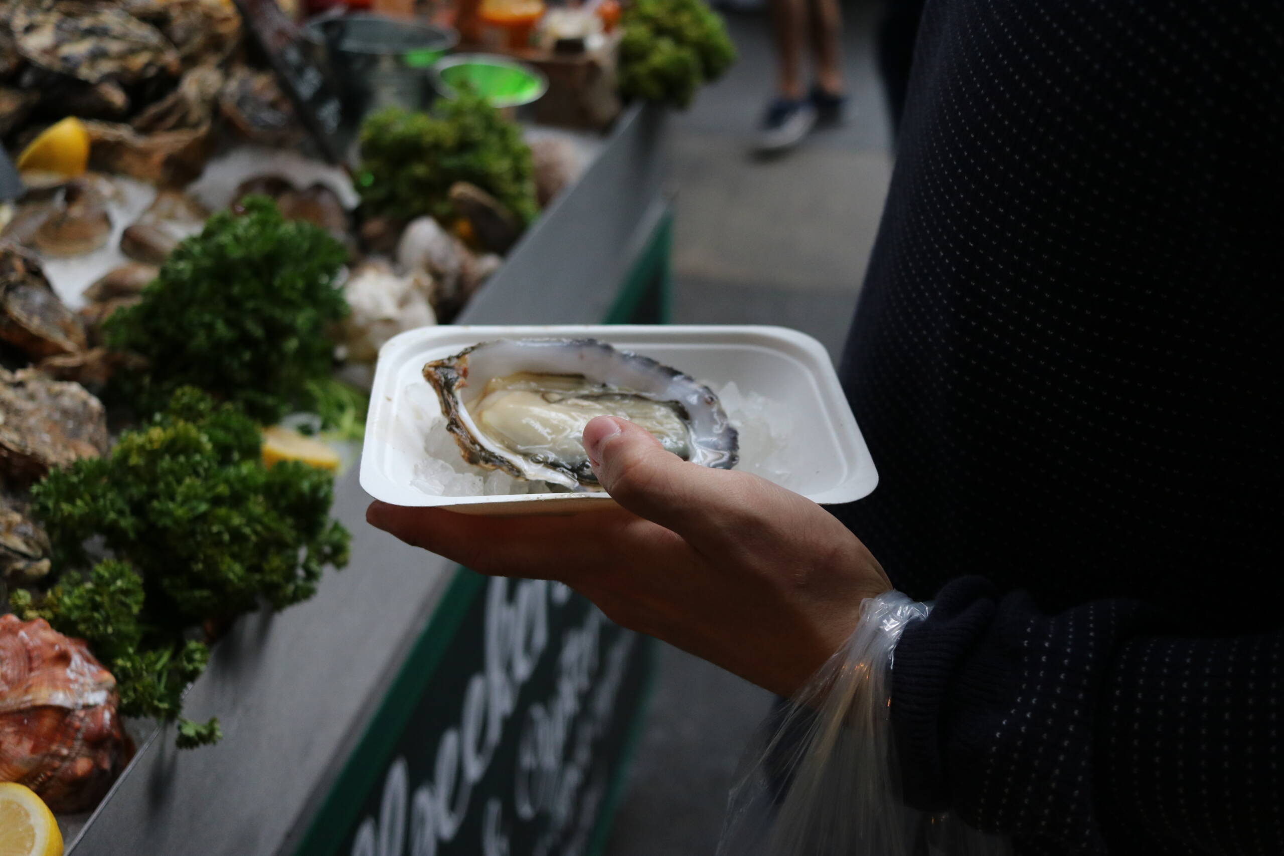 A single shucked oyster on crushed ice in a paper tray, held in one hand at a seafood market stall
