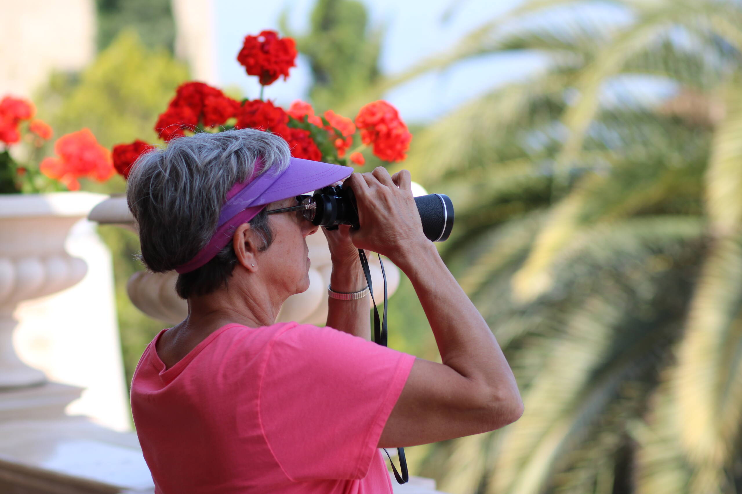 Woman in a pink visor on a flower-lined terrace looking through black roof-prism binoculars, palm trees blurred behind her