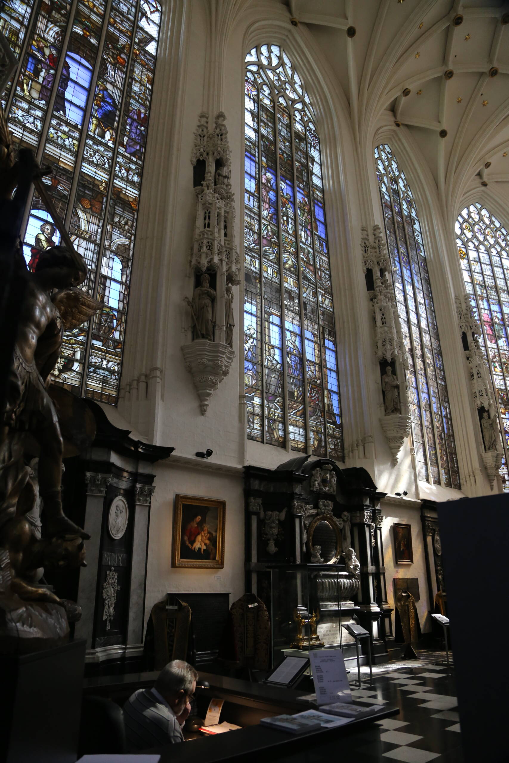 Tall traceried windows filled with narrative stained glass in a whitewashed Brabantine Gothic church interior, with black marble altars and a statue on a carved corbel below