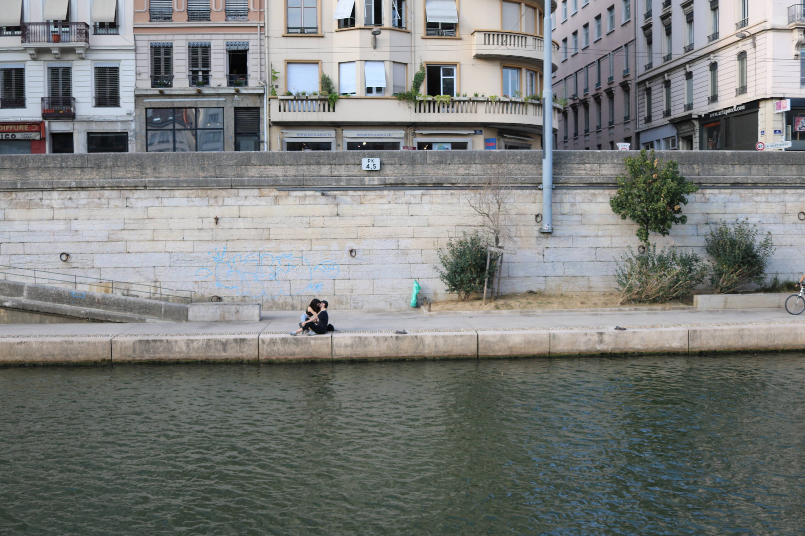A couple sitting on the lower quay of the Saone in Lyon, dwarfed by the ashlar retaining wall and the buildings of Rue de la Martiniere above