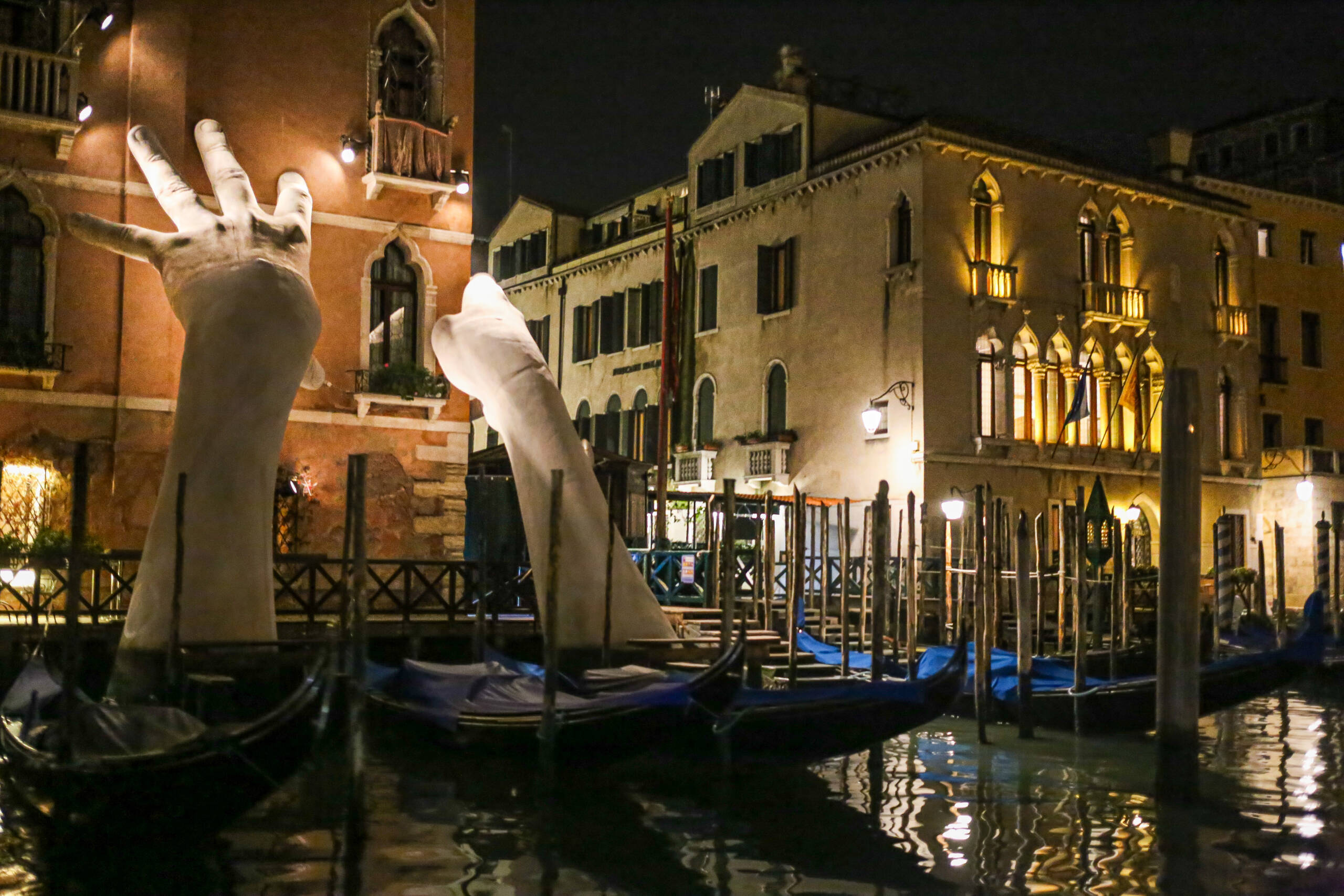 Lorenzo Quinn’s Support lit at night on the Grand Canal, with moored gondolas in the foreground
