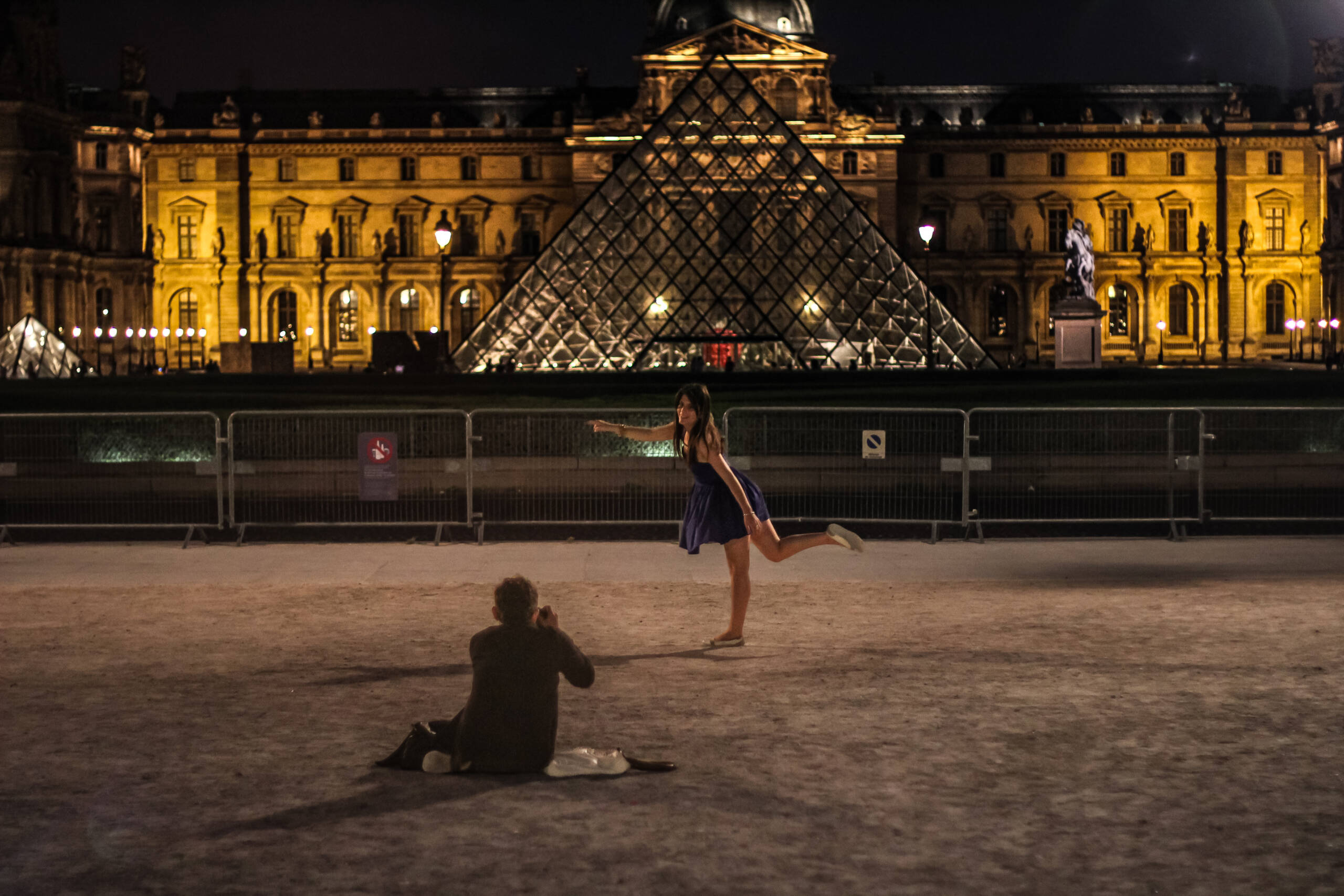 Louvre courtyard at night, a woman posing on one leg while a man sits in the gravel photographing her