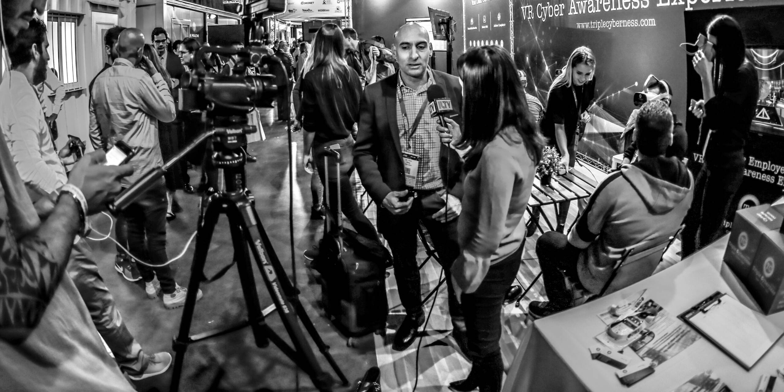 Ultra-wide black-and-white frame of a television interview on a crowded trade show floor, with a camera operator and tripod filling the left foreground