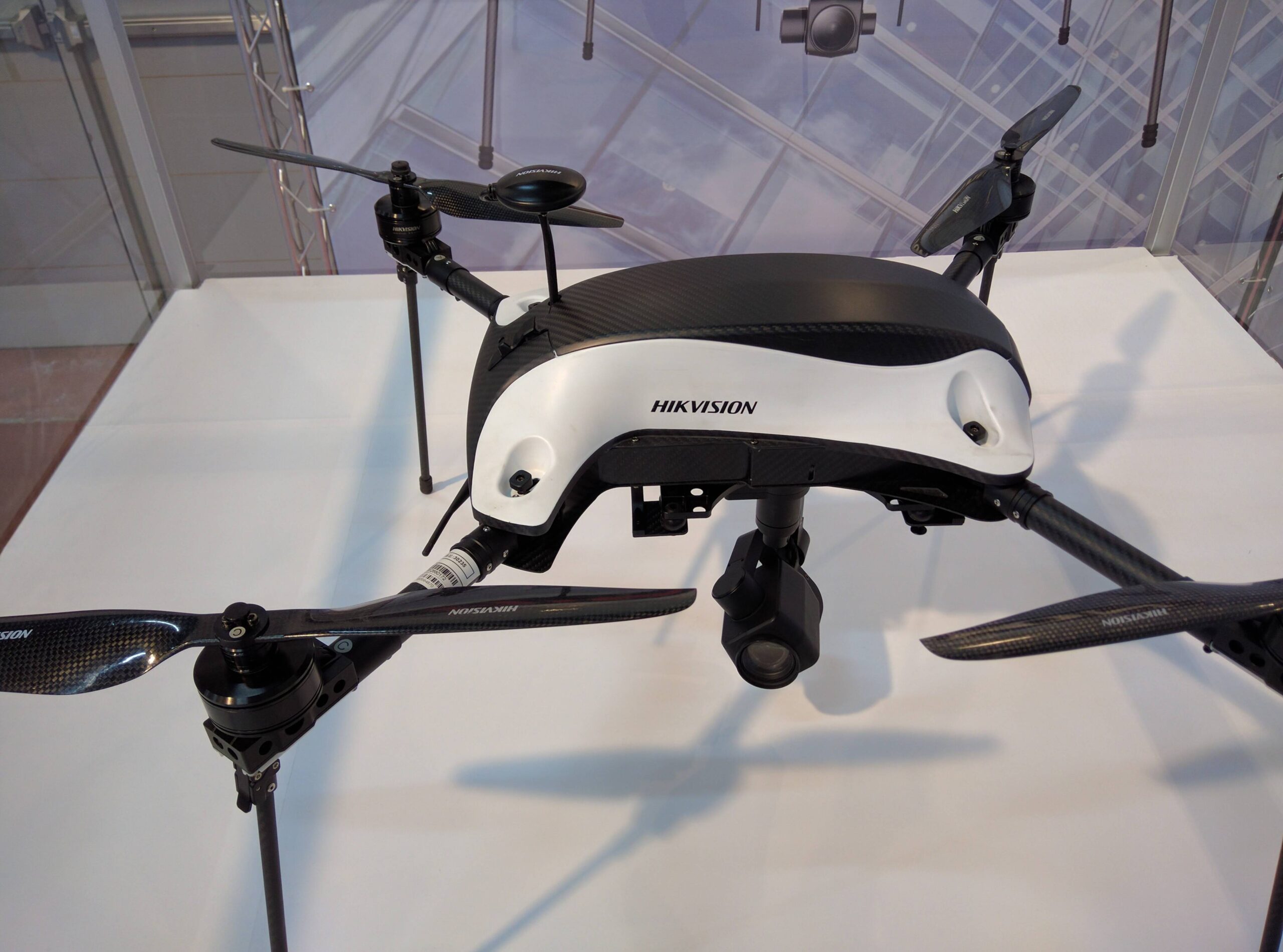 Close view of a black-and-white camera drone on a display table, showing its arms, propellers and hanging camera.