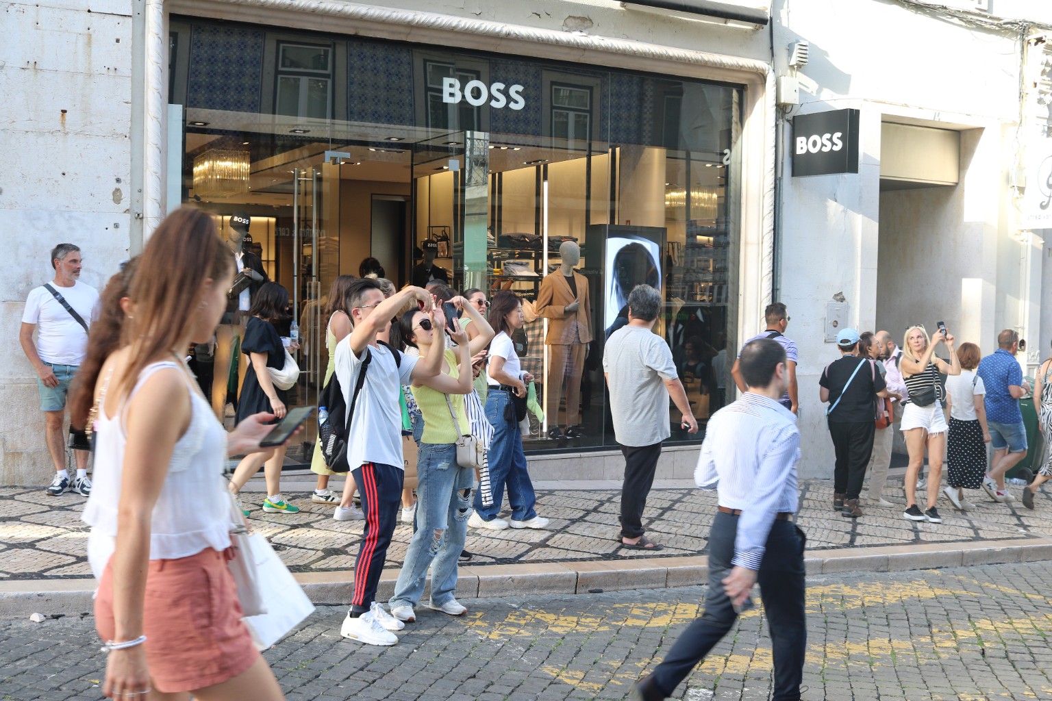 Busy shopping street outside a BOSS store, with a camel-coloured suit in the illuminated window, pedestrians taking photographs, blue tile reflections and patterned stone pavement.