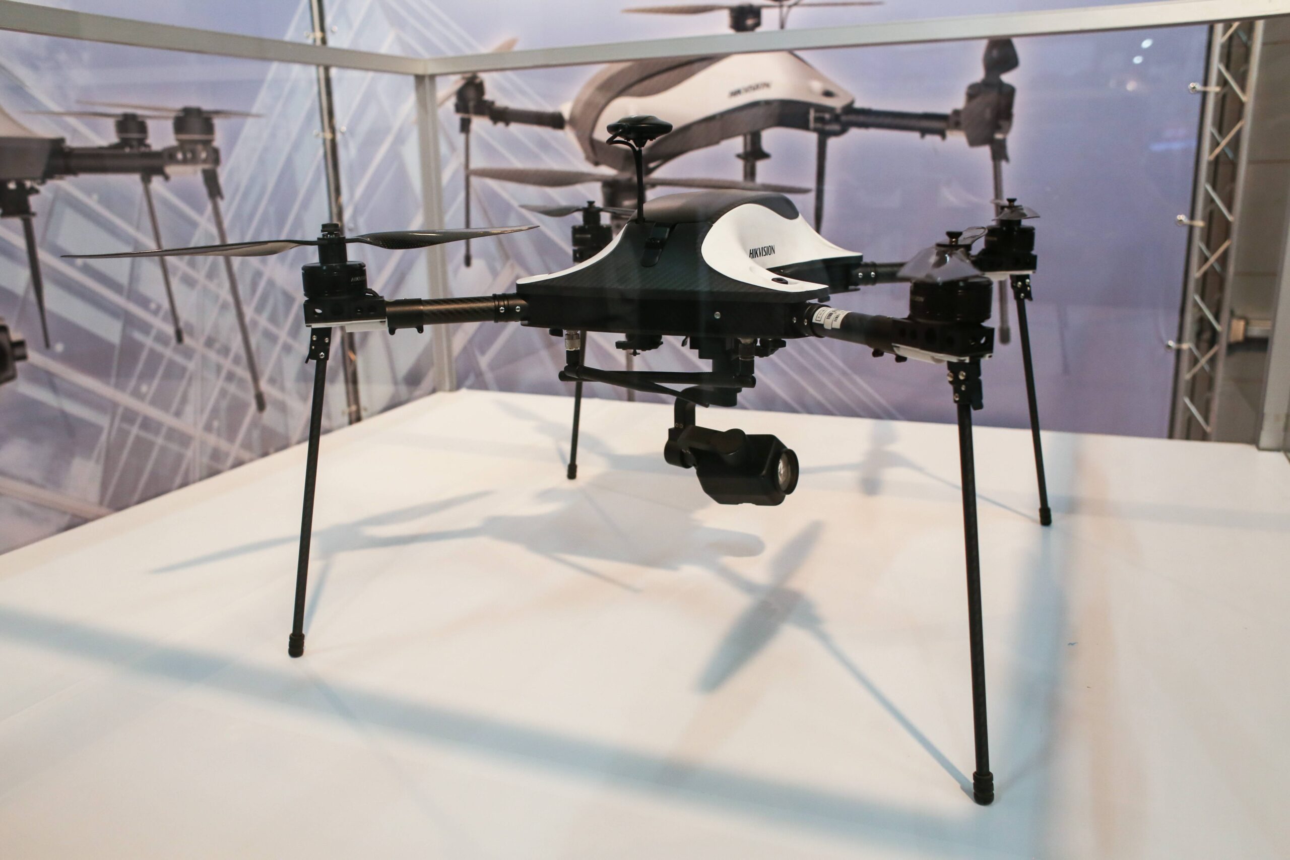 Black-and-white camera drone on tall landing legs at an exhibition, with a suspended gimbal camera and another aircraft behind it.