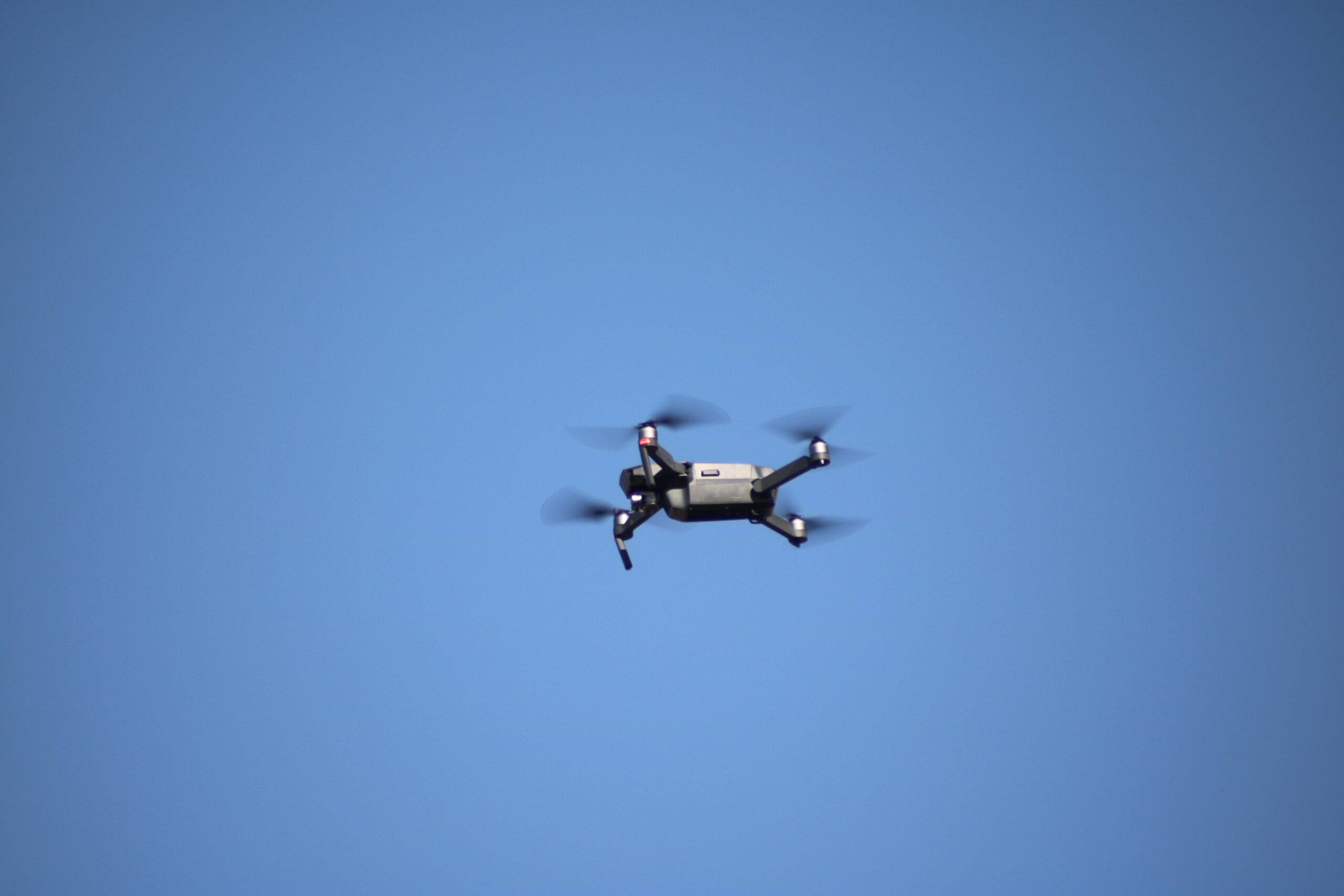 Dark camera quadcopter viewed from below against a blue sky, with four spinning propellers and a suspended camera.