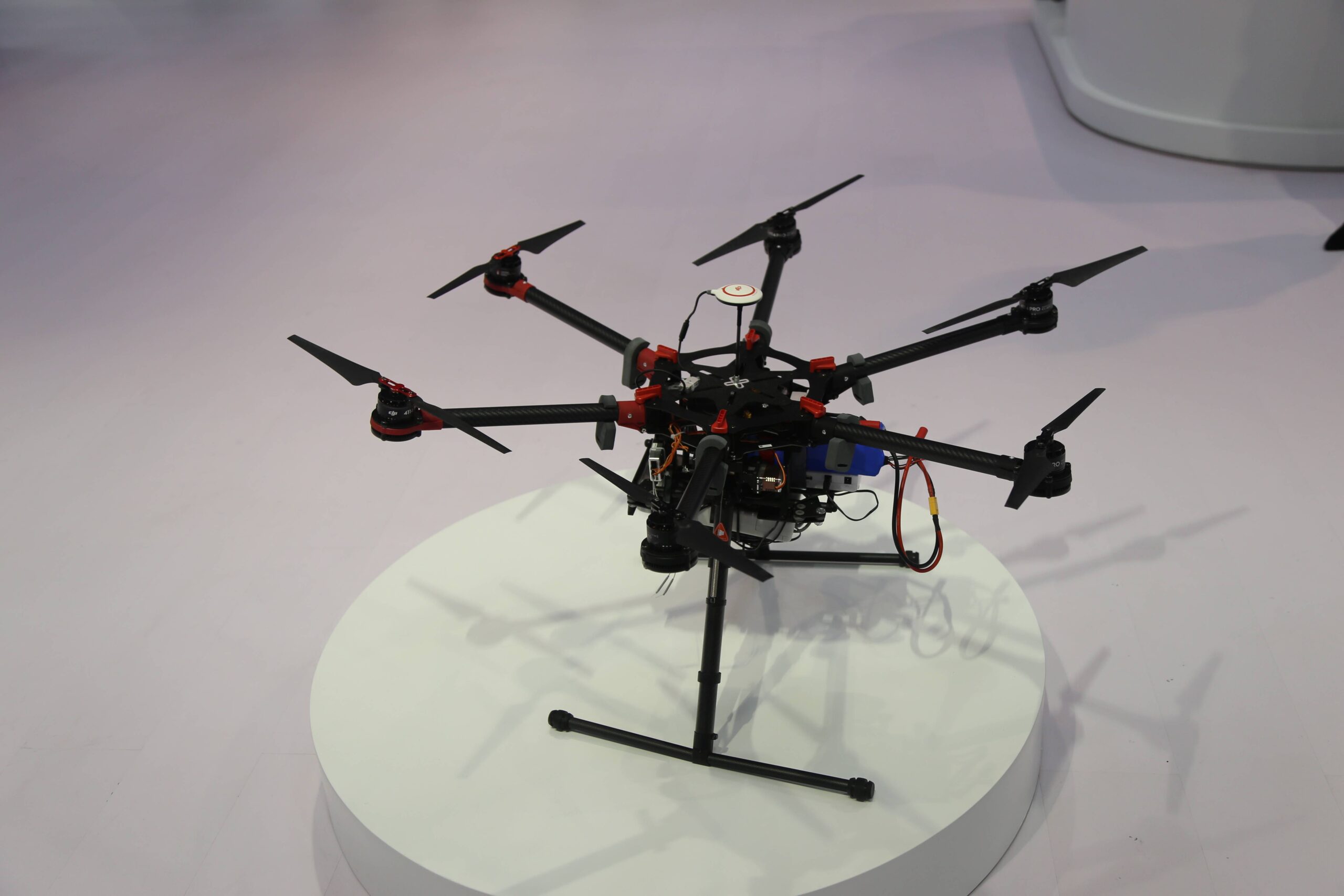 Black exposed-frame multirotor on a round exhibition plinth, with red fittings, visible wiring and a camera payload below.