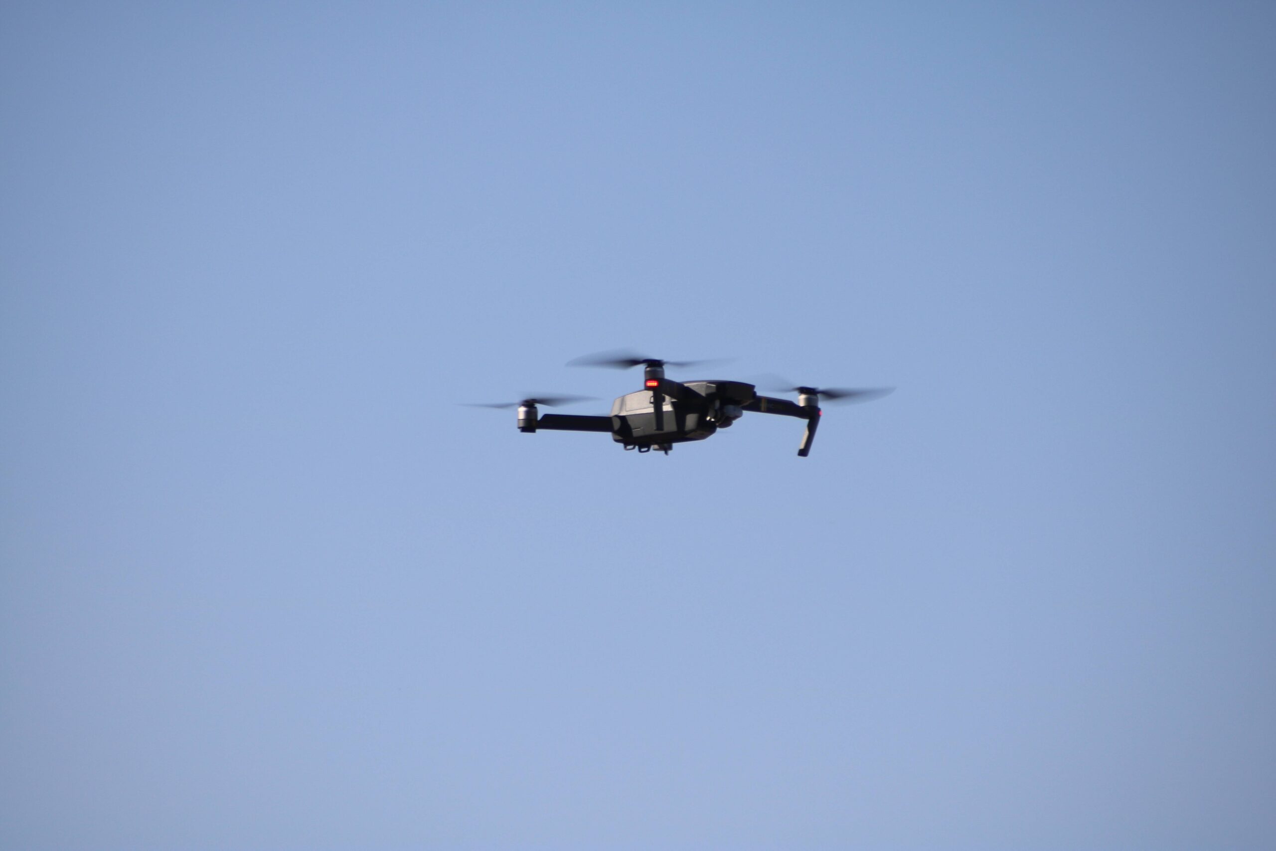 Dark folding quadcopter seen side-on against a clear blue sky, with blurred propellers and a camera beneath the nose.