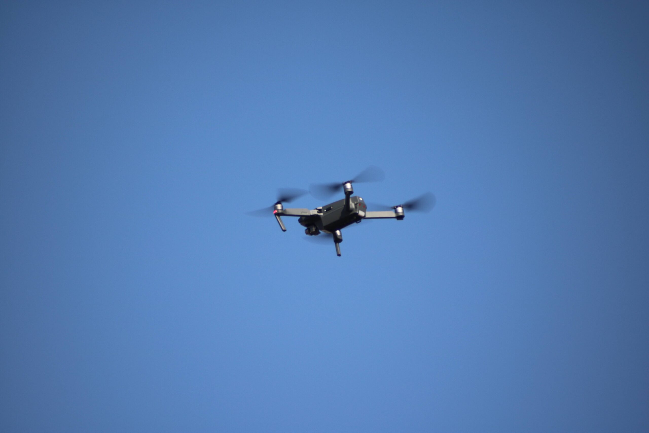 Dark folding quadcopter in flight against a blue sky, with its camera and underside visible.