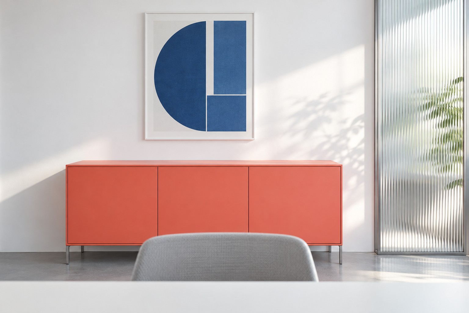 View from webcam height across a desk edge toward a white wall with a low coral cabinet, a blue print and a ribbed glass panel lit by side daylight.