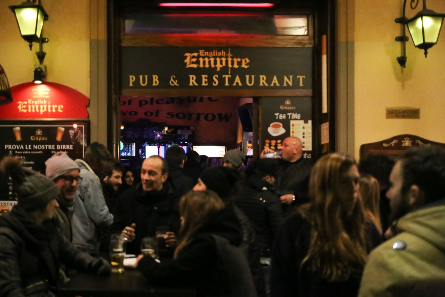 People gathered around outdoor tables outside the English Empire pub at night, beneath yellow wall lanterns and a red light above the entrance.
