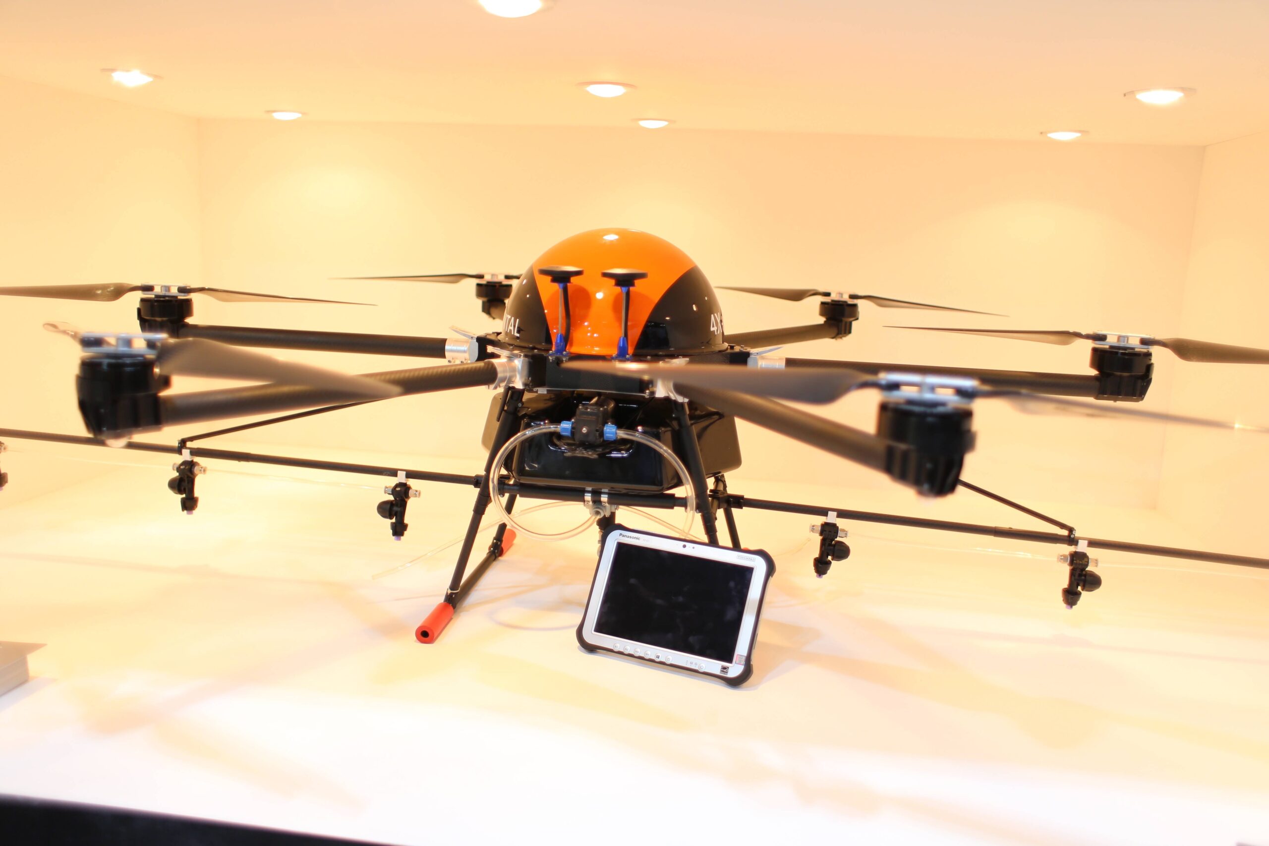 Large orange multirotor aircraft displayed indoors with long arms, landing legs and a small ground screen in front.