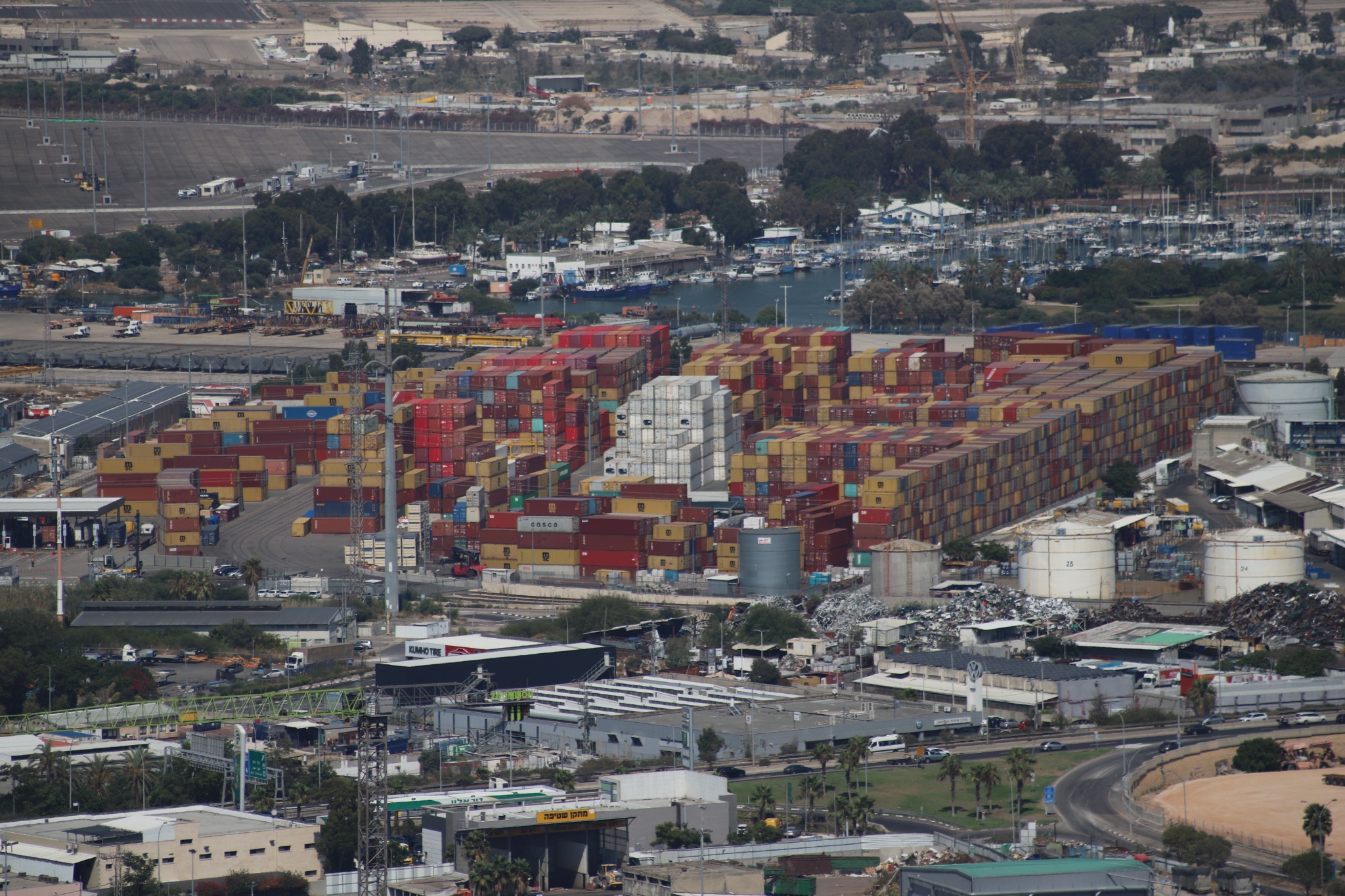 Elevated view of red, yellow, blue and white shipping containers stacked in long rows, with industrial tanks, roads and buildings in front and a marina behind.
