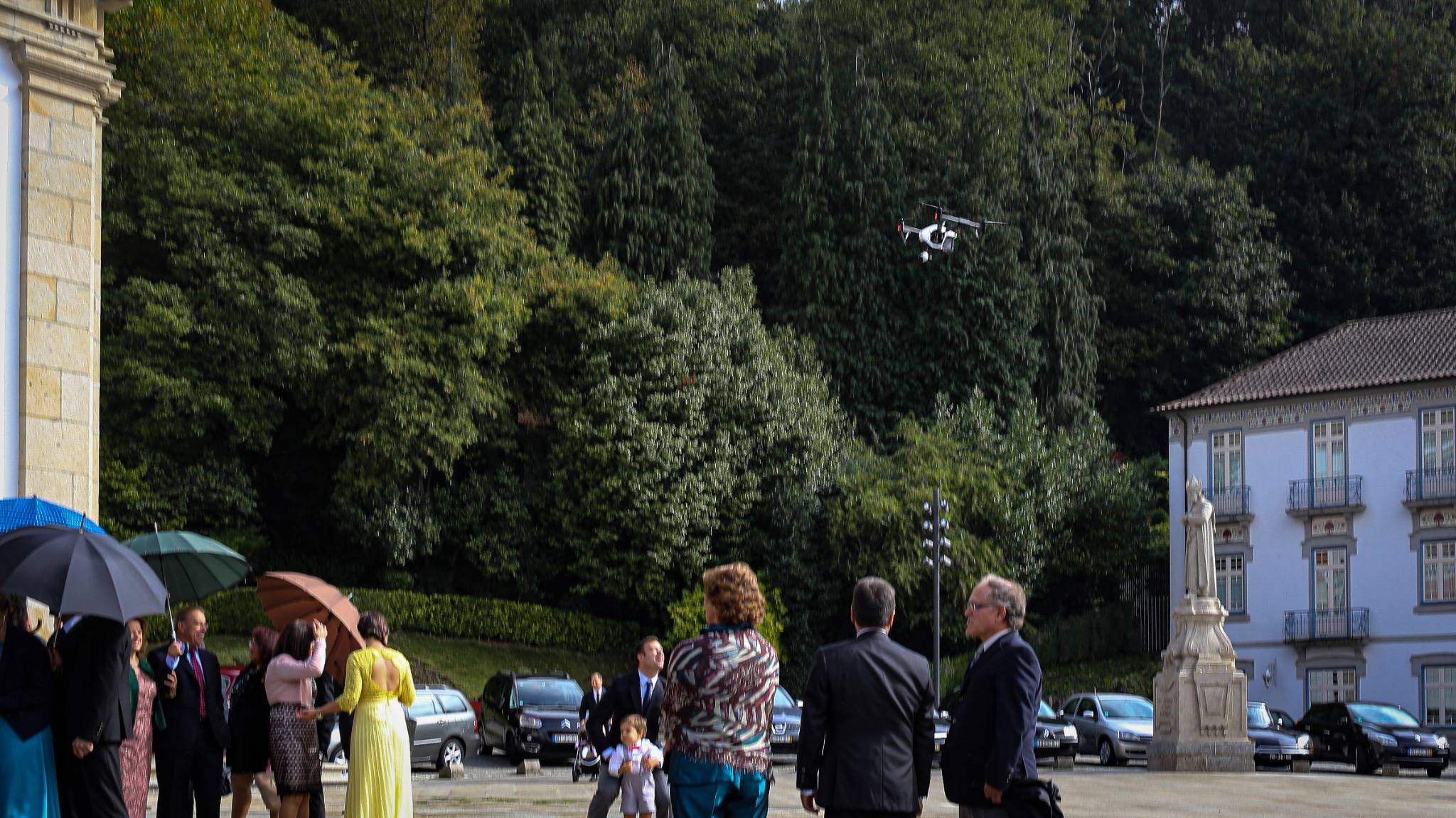 Camera drone in flight above wedding guests with umbrellas in a tree-lined square beside historic buildings.
