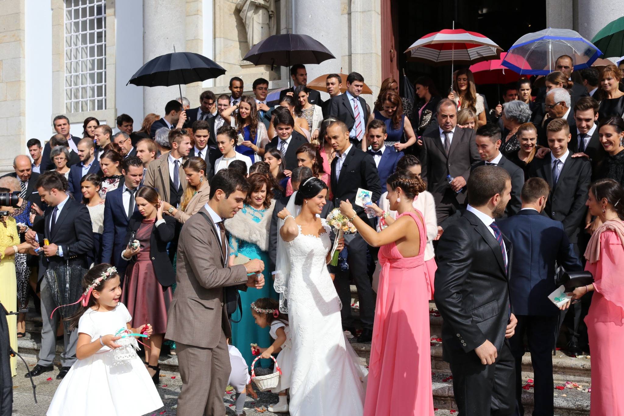 Bride holding a bouquet surrounded by dozens of wedding guests on stone steps, with flower girls, colourful dresses and umbrellas outside a columned entrance.