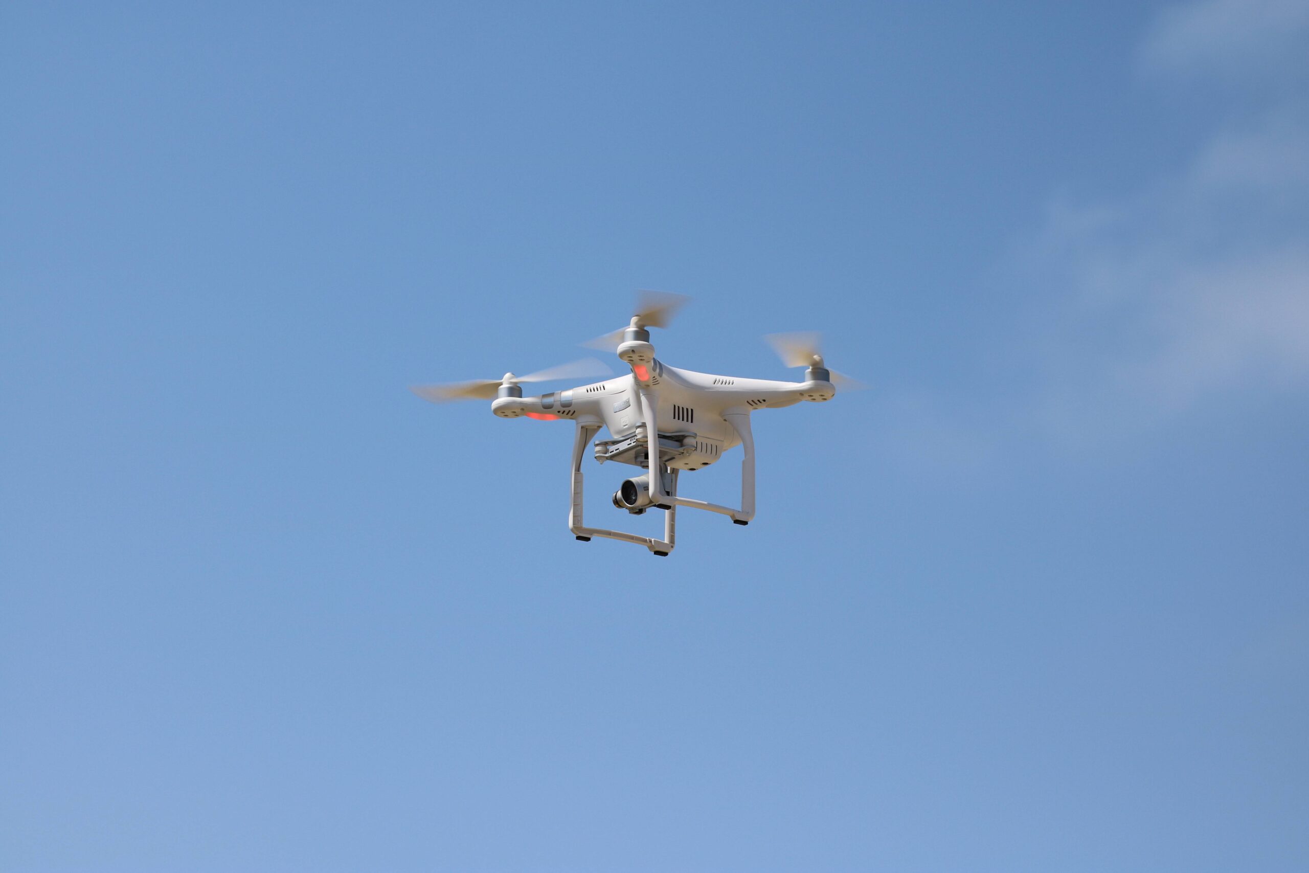 White quadcopter with a camera suspended beneath its body flying against a clear blue sky.